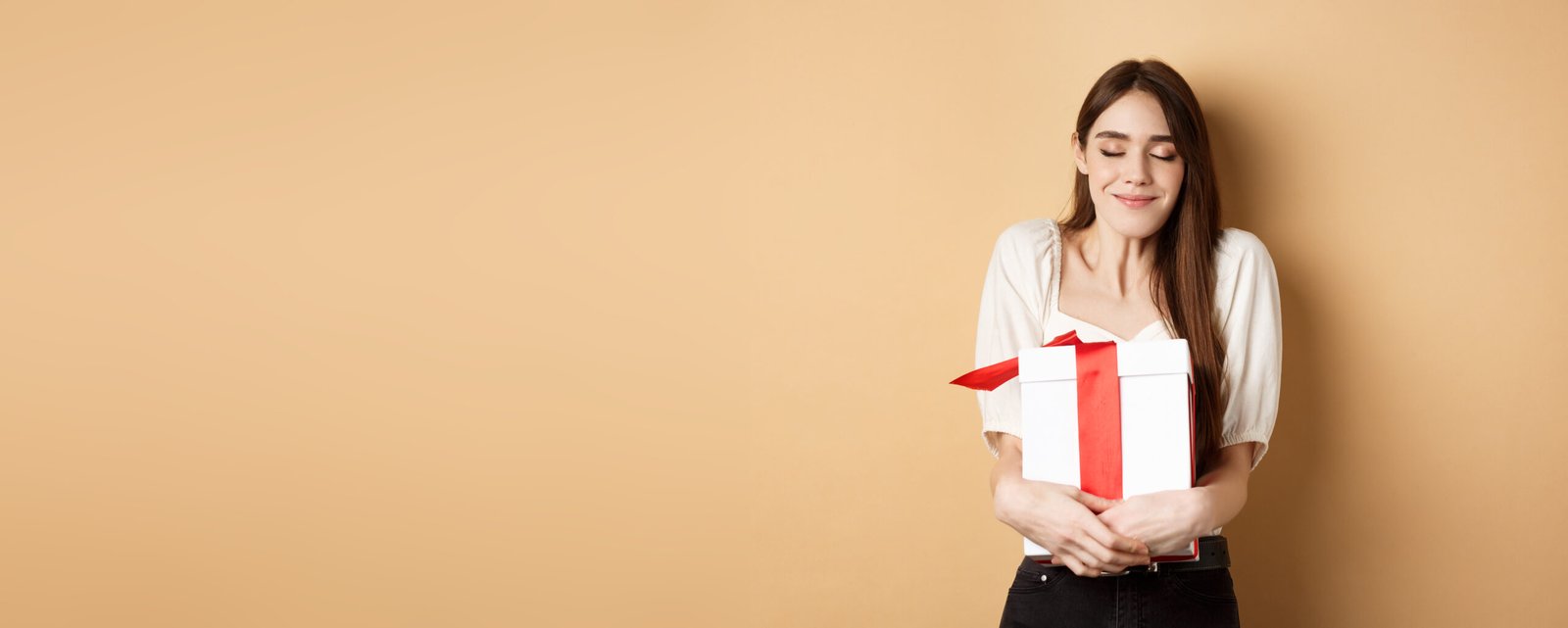 happy valentines day. dreamy woman hugging her surprise gift, close eyes and smile, standing on beige background